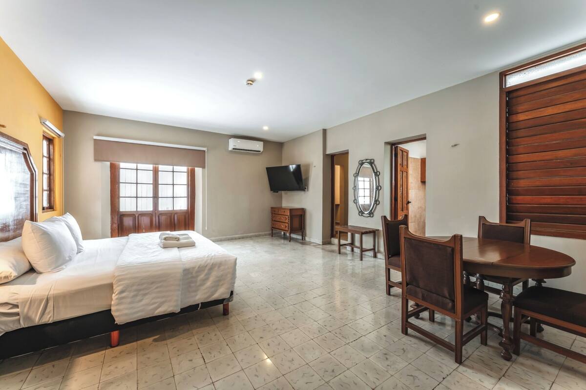 A hotel room with a bed, a TV, a dining table with chairs, and a mirror.