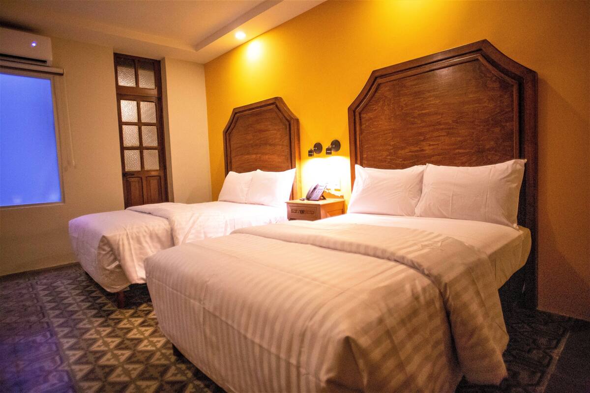 A hotel room with two beds, a wooden headboard, a nightstand with a lamp, and a patterned floor.