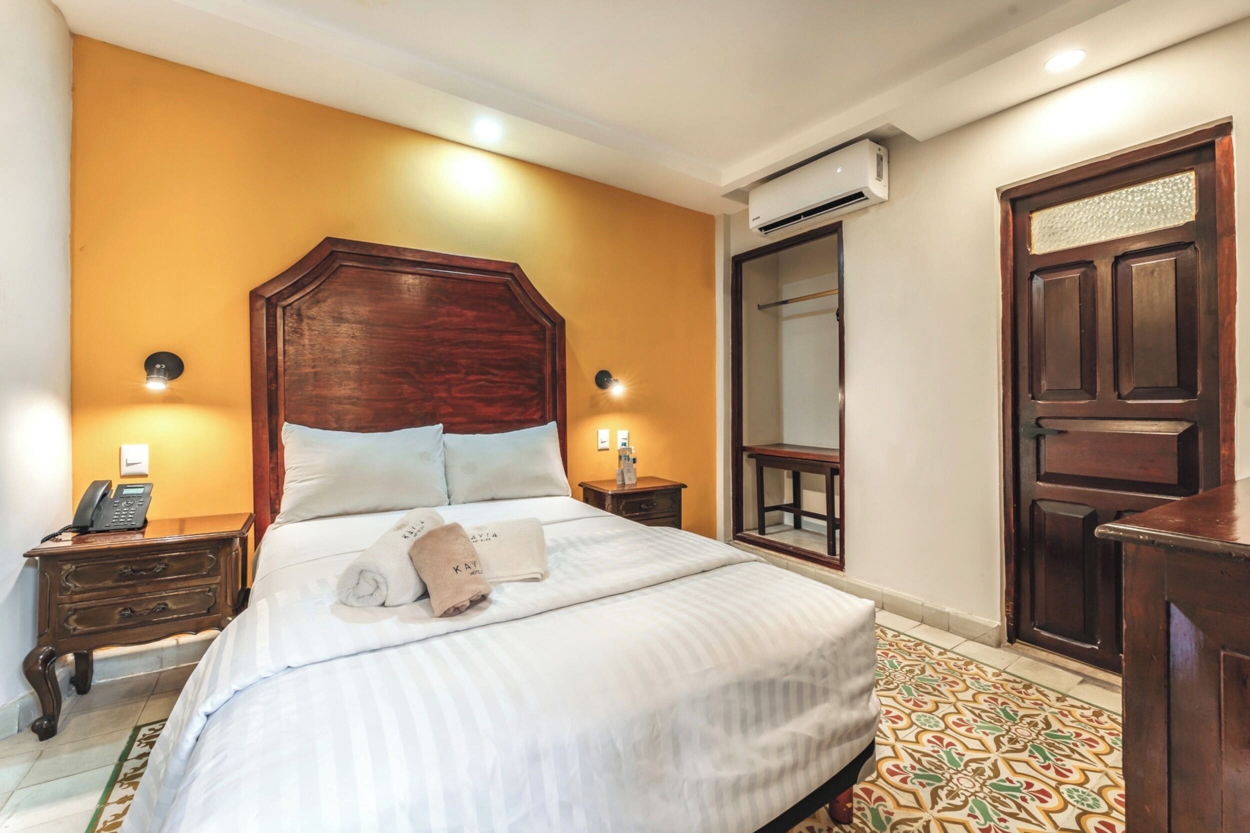 A hotel room with a large bed, wooden headboard, two bedside tables with lamps, a wardrobe, and a patterned floor.