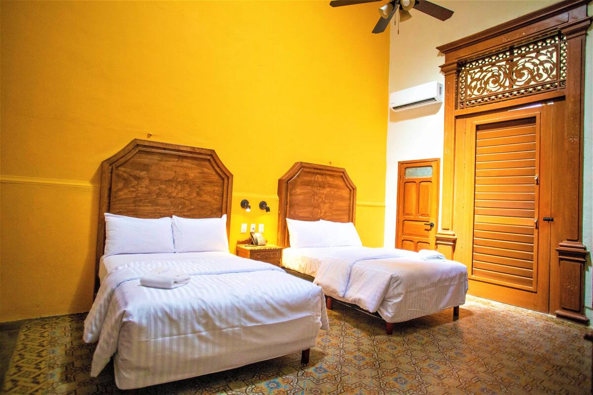 A hotel room with two beds, a ceiling fan, an air conditioning unit, and a wooden wardrobe.