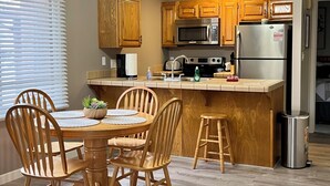 A modern kitchen with wooden cabinets, a stainless steel refrigerator, a microwave, and a sink. There is a dining table with chairs and a small stool.
