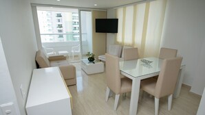 A modern living room with a glass dining table, white sofas, and a flat-screen TV mounted on the wall. - 2TC10-J APARTAMENTO FRENTE AL MAR (Cartagena)