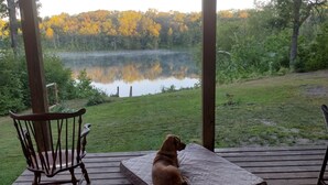 Eine Holzterrasse mit einem Hund, der auf einer Matratze sitzt und auf einen See mit Bäumen im Hintergrund blickt.