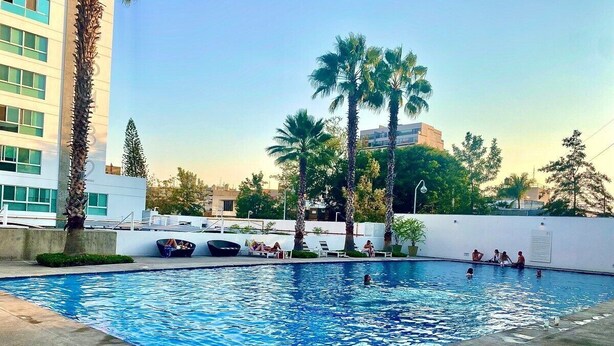 A swimming pool with people around it, palm trees, and a building in the background. - 2 BEDROOMS WITH POOL, GYM, 24 HOURS SECURITY, 7 PEOPLE (Guadalajara)