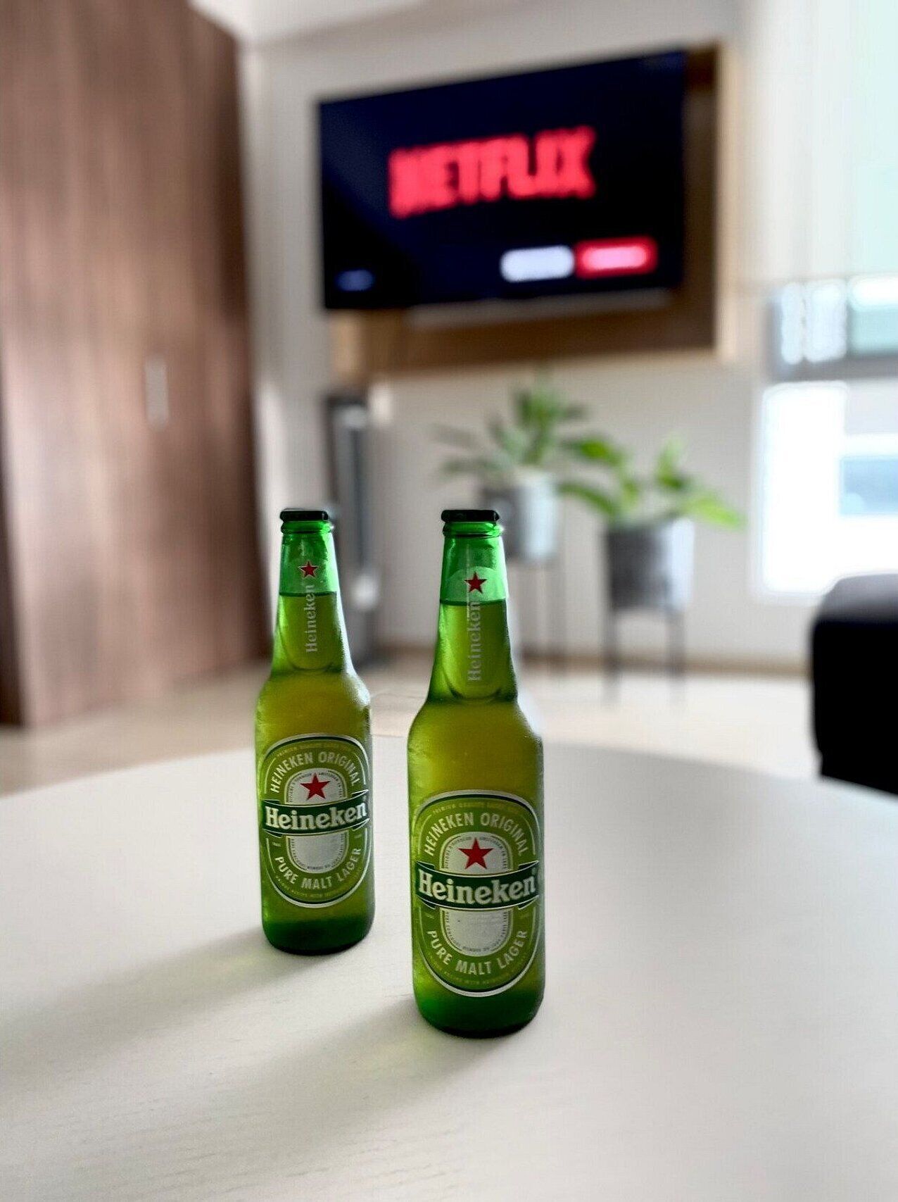 Two Heineken beer bottles on a table in a room with a TV displaying Netflix in the background.