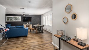 Uma sala de estar moderna com um sofá azul, mesa de jantar de madeira e uma TV fixada numa parede de tijolos.