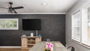 Uma sala de jantar com mesa de madeira, cadeiras, ventilador de teto, TV de tela plana fixada em uma parede de tijolos e uma janela com persianas.