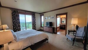 A hotel room with a large bed, a desk with a computer, a chair, and a TV. There are curtains on the windows, and a balcony is visible through the door. - Executive House Suite with Gorgeous Olympic Views (Port Angeles)