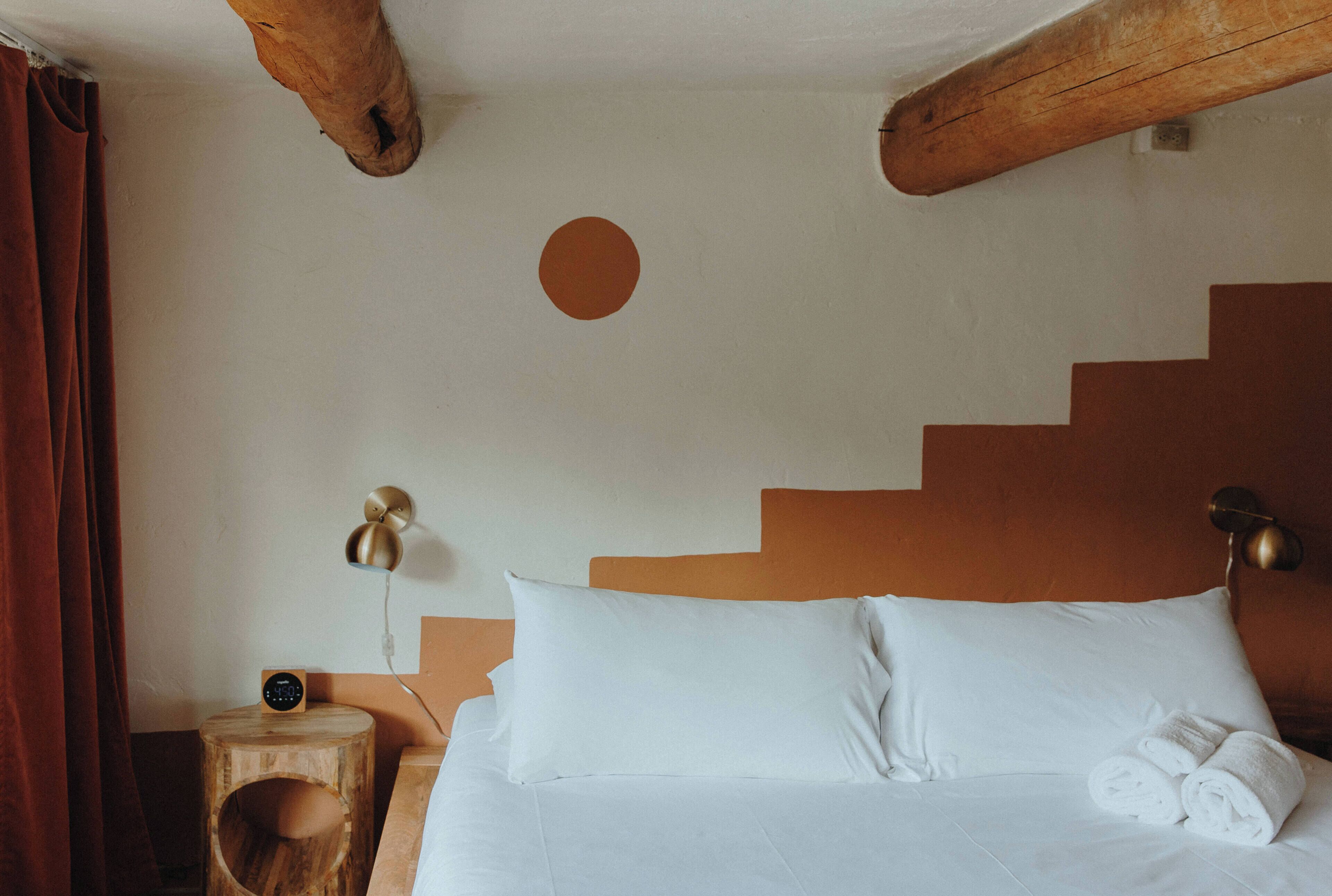 Una cama con sábanas blancas, una mesita de noche de madera con una lámpara, y una pared con un recorte circular.