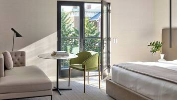 A hotel room with a bed, a green chair, a small table with books, and a balcony with a view of buildings and trees.