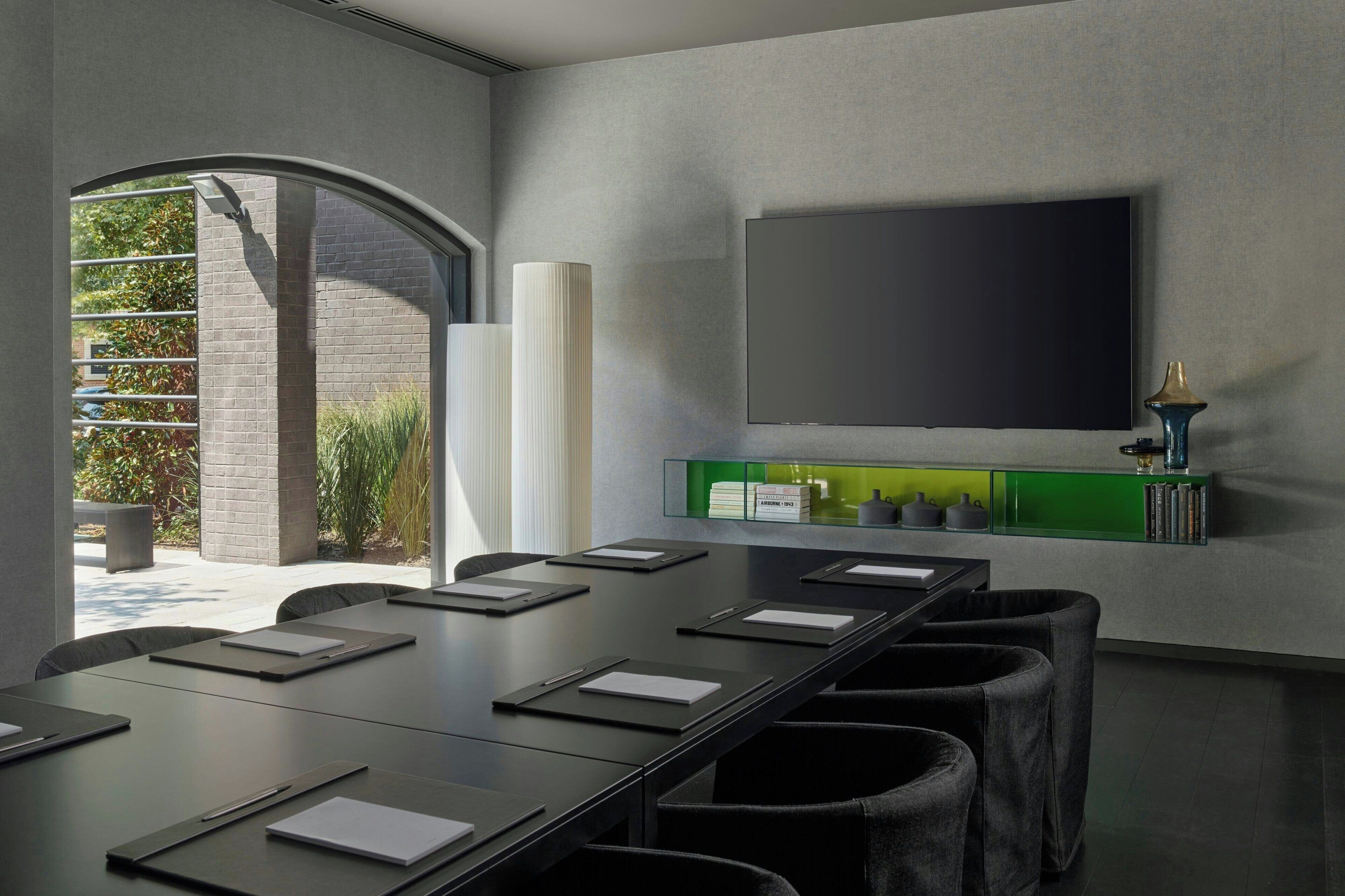 Un salón de conferencias con una larga mesa, sillas, un televisor montado y un estante verde con objetos.