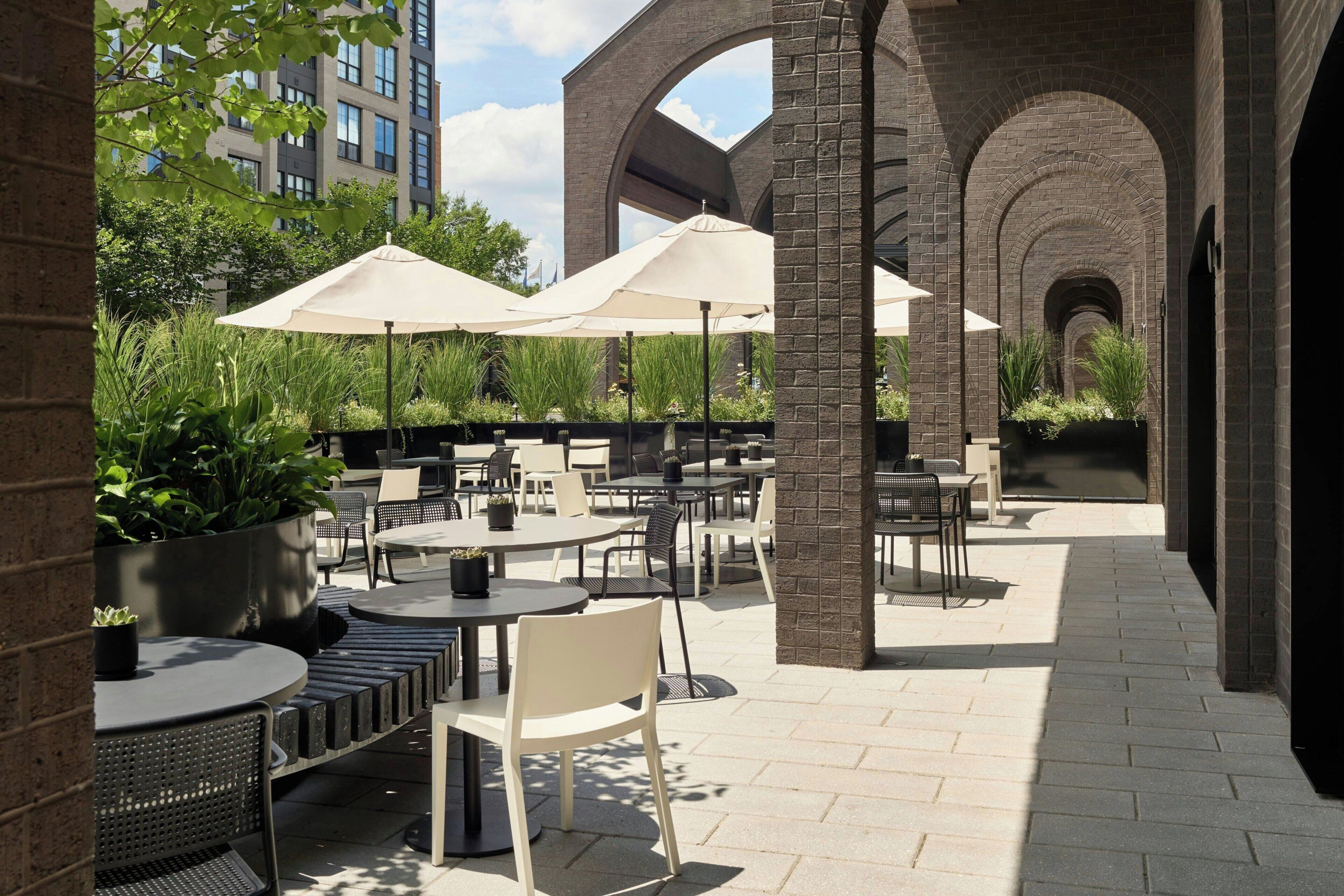 An outdoor patio area with tables and chairs, surrounded by brick arches, and potted plants.