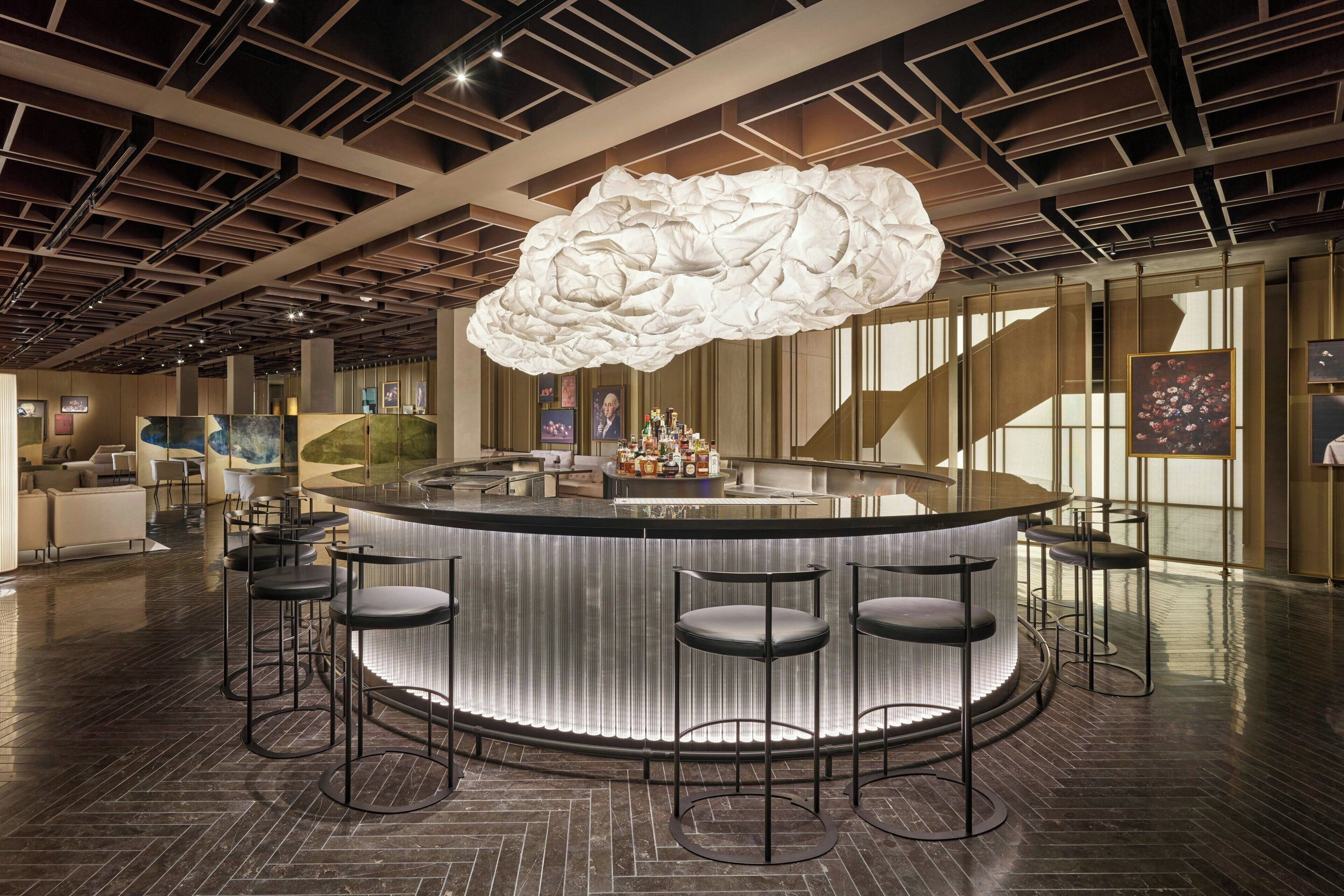 A modern bar area with a unique cloud-shaped light fixture, bar stools, and a polished wooden floor.