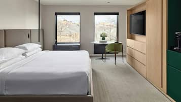 Ein Hotelzimmer mit einem großen Bett, einem Fernseher, einem Schreibtisch mit Stuhl und Blick auf Gebäude durch die Fenster.