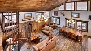 Ein geräumiger Wohnbereich mit Holzboden, einer Holztreppe, einem Kamin, einer Couch, einem Esstisch mit Stühlen und großen Fenstern mit Jalousien.
