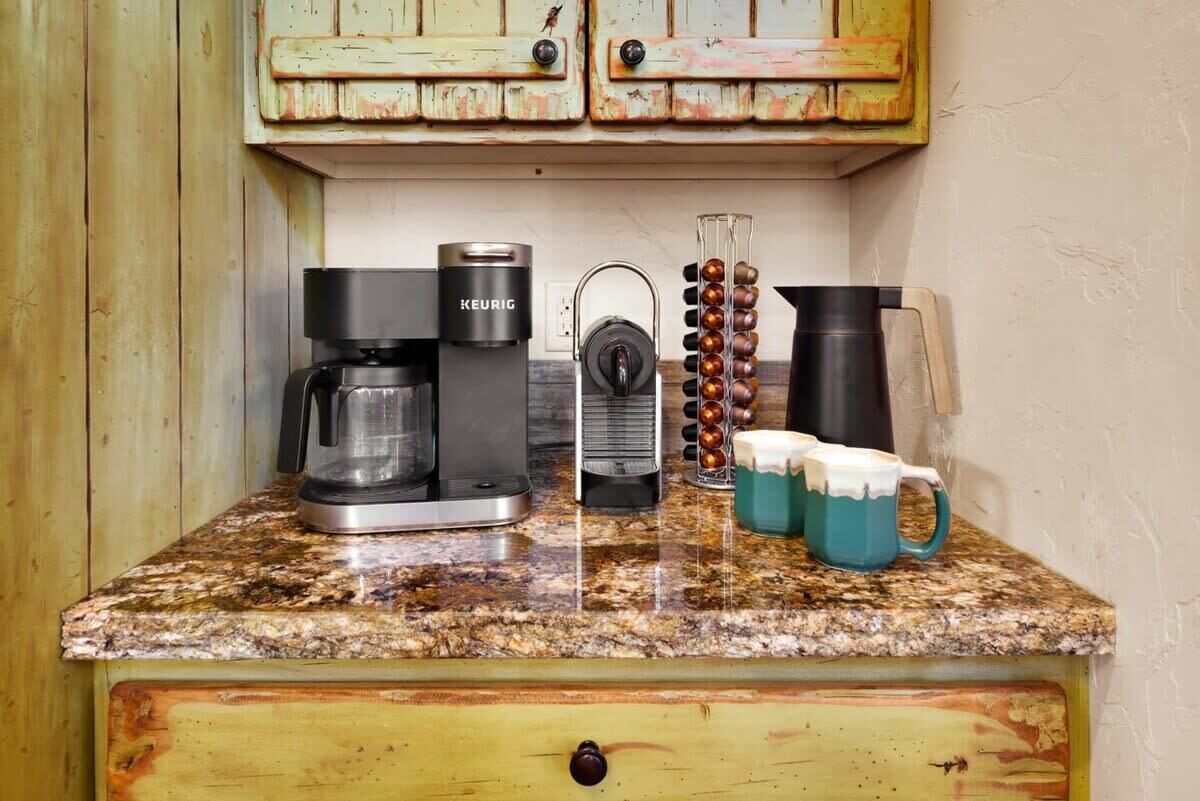 A kitchen counter with a coffee maker, a kettle, a coffee grinder, and two mugs.