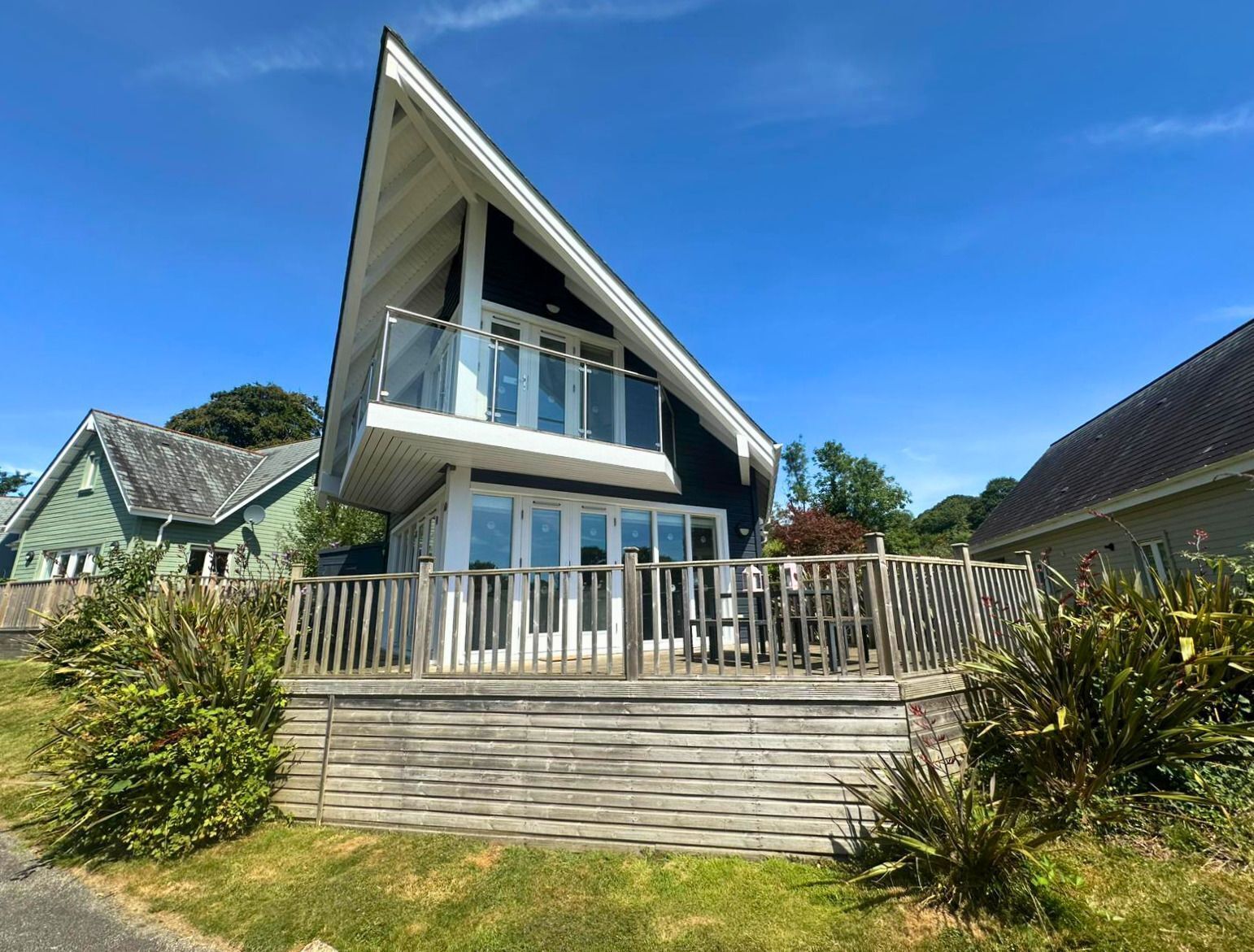 A modern two-story house with a balcony, surrounded by a wooden deck and greenery.