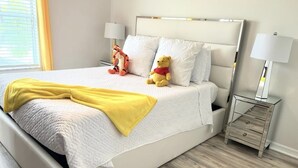A neatly made bed with a white comforter and a yellow throw blanket, flanked by two plush toys. A bedside table with a lamp is visible to the right.