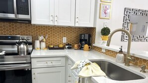 A kitchen with white cabinets, a stainless steel microwave, a kettle, and a sink with a gold faucet.
