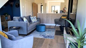 A modern living room with a grey sofa, a blue ottoman, and a grey armchair. There is a wooden floor and a large window in the background. - Porth Oer, luxury accommodation (Nefyn)