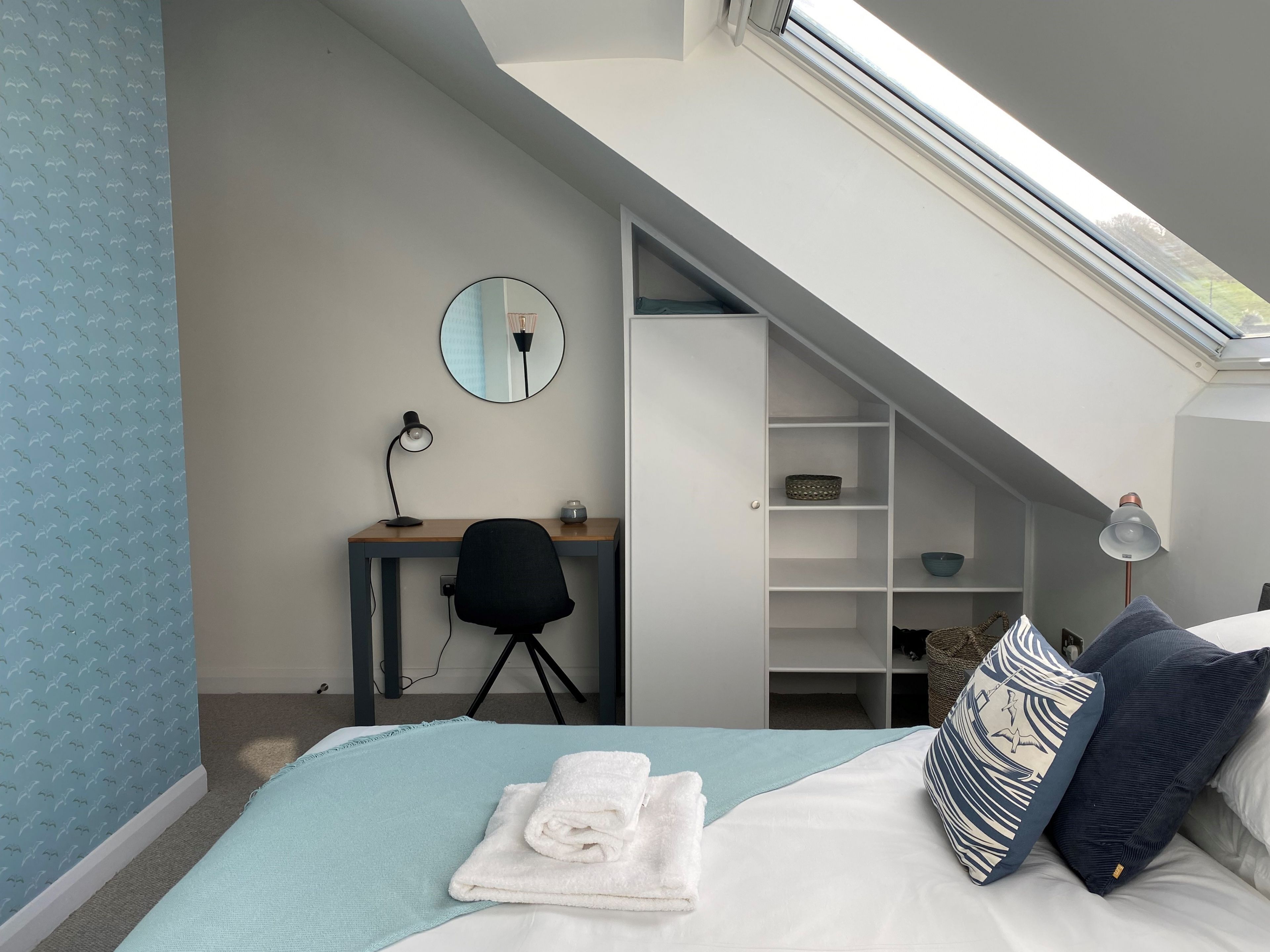 A bedroom with a bed, a desk, a chair, a mirror, a skylight, and a built-in closet.