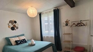 A bedroom with a bed, a bedside table, a hanging light fixture, a window with curtains, and a metal shelf with hangers. - Detached house with enclosed garden (Abbeville)