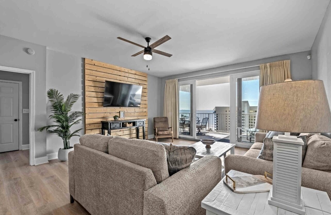 A modern living room with a sofa set, a TV mounted on a wooden panel wall, a ceiling fan, and a balcony with a view.
