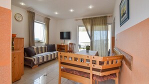 A cozy living room with a sofa, a wooden dining table with chairs, a television, and a large window with curtains. - Apartments Jaklin - Two Bedroom Apartment with Terrace and Sea View (Rab)
