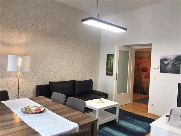 Een moderne kamer met een houten eettafel, een bank, een witte bijzettafel en een lamp. Een deur leidt naar een andere kamer met een schilderij en een rode bloem.