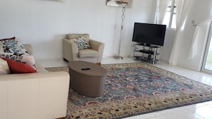 A living room with a beige sofa, a smaller sofa with patterned cushions, a floor lamp, a flat-screen TV on a stand, and a patterned rug. - 5 Mins from beach! (Mero)