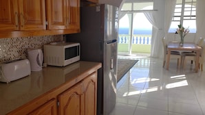 A kitchen with wooden cabinets, a microwave, a toaster, and a refrigerator. There is a dining area with a table and chairs, and a view of the sea through a glass door. - 5 Mins from beach! (Mero)