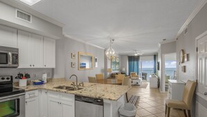 Una cocina moderna con armarios blancos, encimera de granito y lavavajillas. Hay un comedor con mesa y sillas, y vistas al océano a través de amplias ventanas.