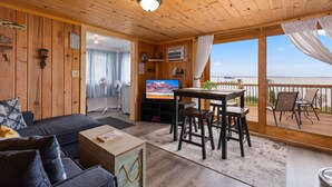 A cozy cabin interior with a wooden ceiling, a TV, a dining area, and a balcony with a view of the water. - Cozy cabin steps from the lake! (Brainerd)