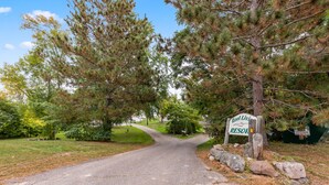 A sign for Reel Living Resort with a paved driveway leading through a wooded area. - Cozy cabin steps from the lake! (Brainerd)