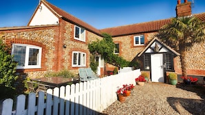 A stone house with a white picket fence, a small garden, and a door with a window. - Edelweiss | East Ruston Cottages (Happisburgh)