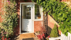 A brick house with a white door, a small porch, and a garden with potted plants and a bench. - Edelweiss | East Ruston Cottages (Happisburgh)