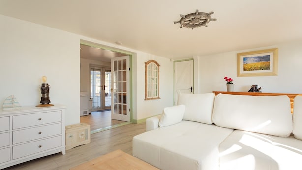 A spacious living room with a white sofa, wooden coffee table, and a white dresser. A door leads to another room, and a framed picture hangs on the wall. - Edelweiss | East Ruston Cottages (Happisburgh)