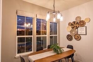 Uma área de refeições com mesa de madeira, cadeiras, luminária pendente e vista do céu através de grandes janelas.