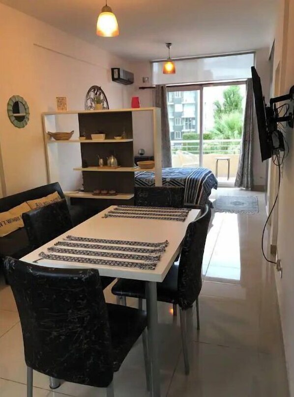 Un salon moderne comprenant un coin repas, une table blanche agrémentée de chemins de table noirs et blancs, des chaises noires, une étagère blanche ornée d’objets décoratifs et une grande fenêtre donnant sur un paysage verdoyant.