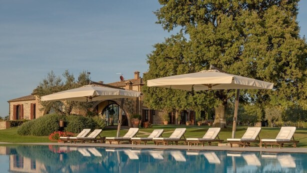 Un área de piscina con sillones de descanso y sombrillas, rodeada por un edificio de piedra y árboles.