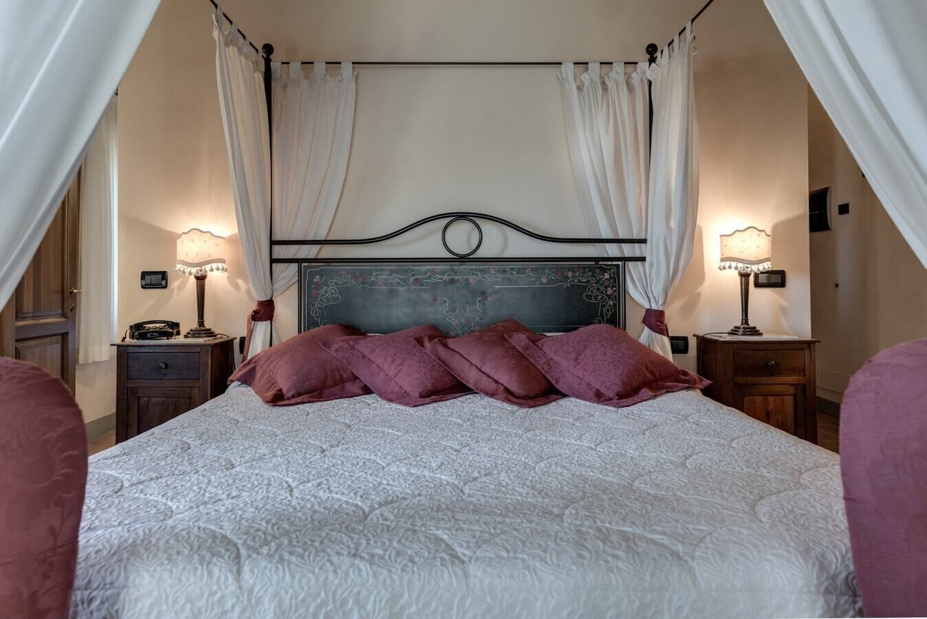 A four-poster bed with a white quilt and purple pillows, flanked by two bedside tables with lamps, in a room with cream walls and a wooden floor.