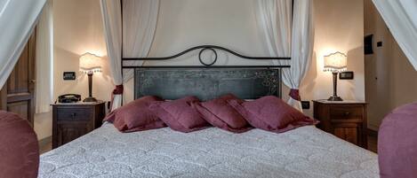 A four-poster bed with a white quilt and purple pillows, flanked by two bedside tables with lamps, in a room with cream walls and a wooden floor.