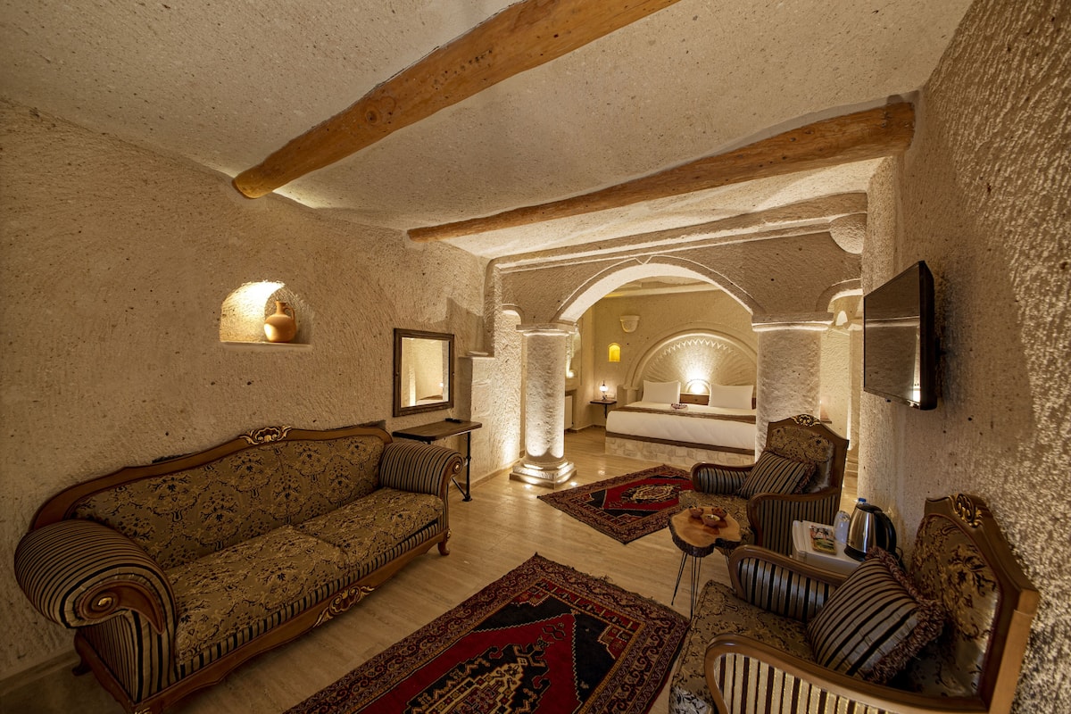 A room with a sofa, armchairs, a coffee table, and a fireplace. There is a large pillar and a bed in the background.