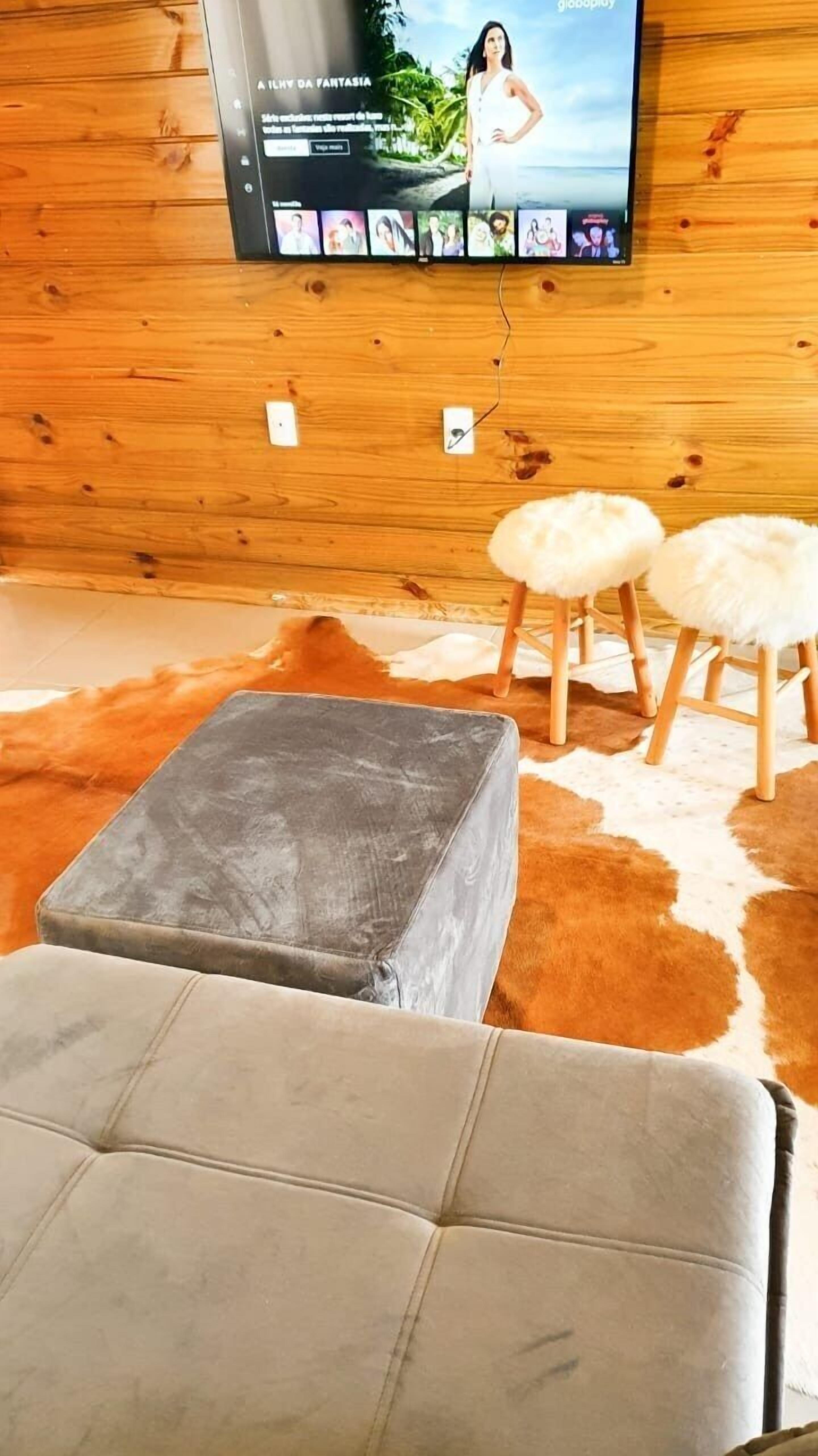 Ein Wohnzimmer mit einem grauen Hocker, zwei weißen, fellartigen Hockern, einem Holzboden und einem an der Wand montierten Fernseher, der eine Frau zeigt.