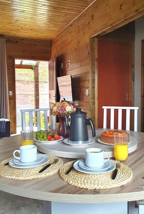 Una mesa de comedor de madera con desayuno, incluyendo café, jugo y un pastel, ubicada en una veranda con vista a follaje verde.