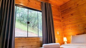 Quarto de cabana de madeira com uma cama, uma janela com cortinas e vista para uma floresta.