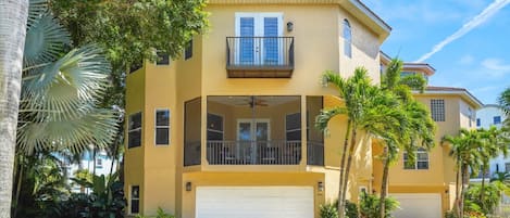 A two-story yellow building with a white garage door, surrounded by palm trees and greenery.