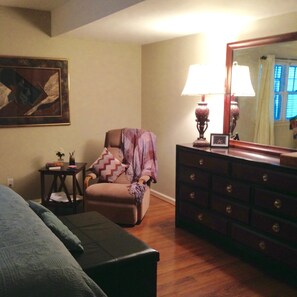 Une pièce avec un plancher en bois, une chaise recouverte d’une couverture, une commode à tiroirs, une lampe et un tableau accroché au mur.