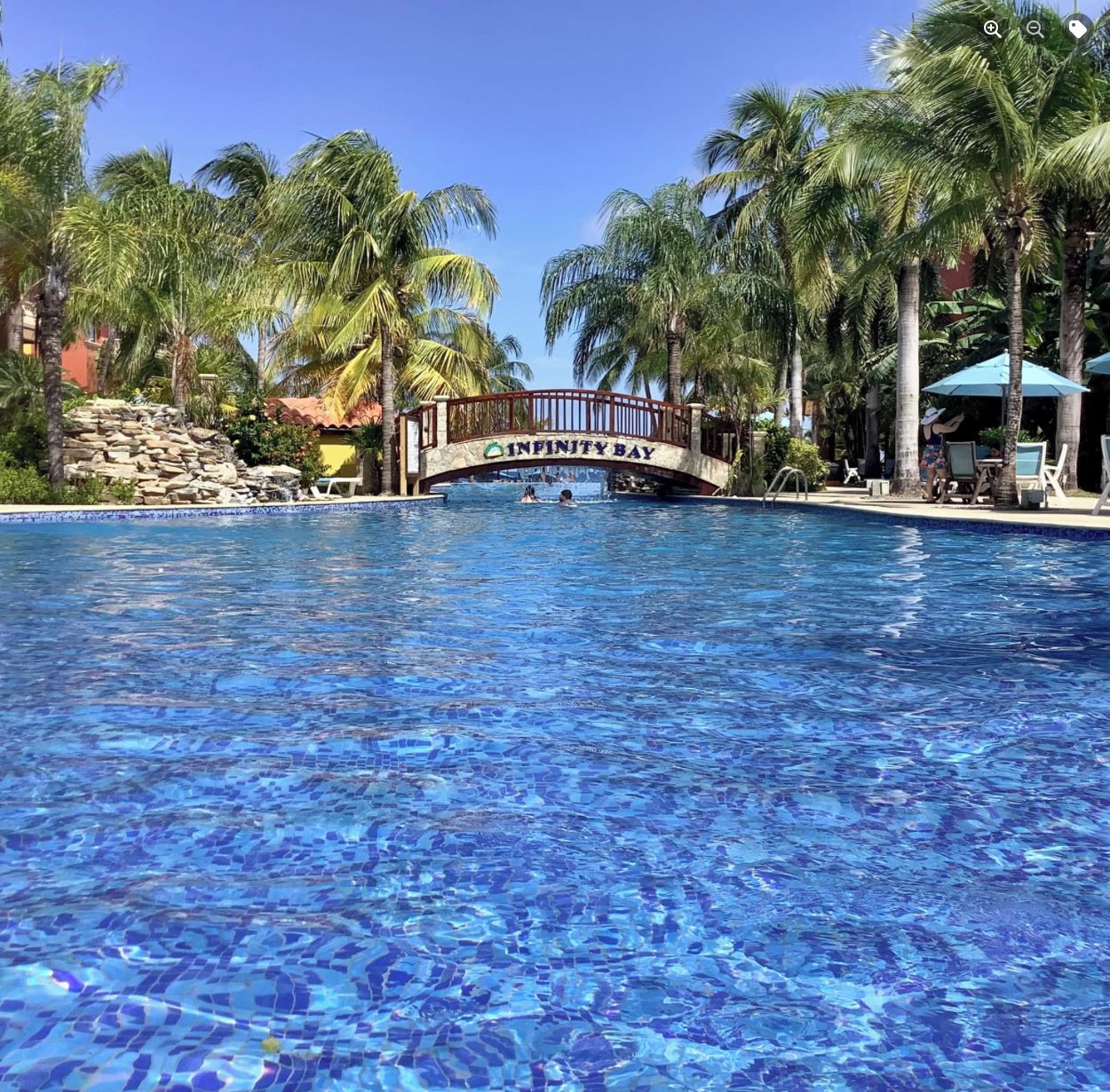 Ein Swimmingpool mit einer Brücke, auf der „Infinity Bay“ steht, umgeben von Palmen und Liegestühlen.