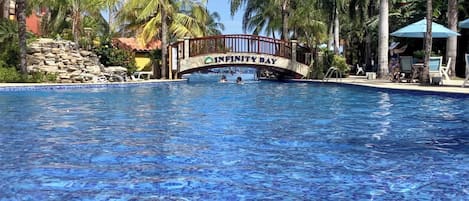 Ein Swimmingpool mit einer Brücke, auf der „Infinity Bay“ steht, umgeben von Palmen und Liegestühlen.